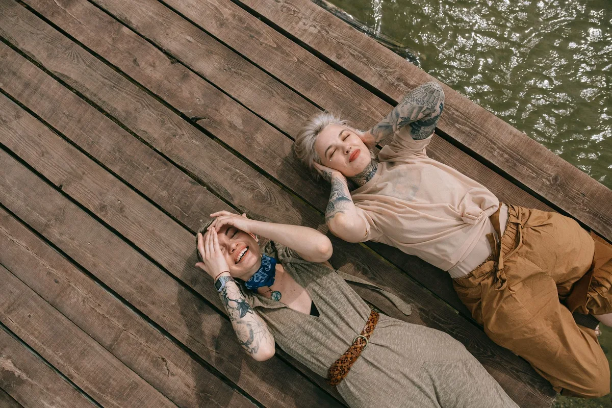 Two cheerful friends with tattoos lying on a wooden pier by the water, enjoying sunlight.