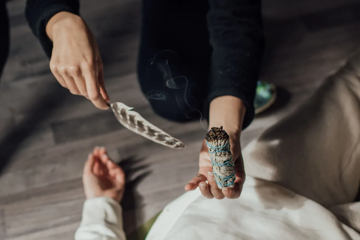 Hands using a feather and smudge stick in a spiritual ritual, symbolizing purification and meditation.
